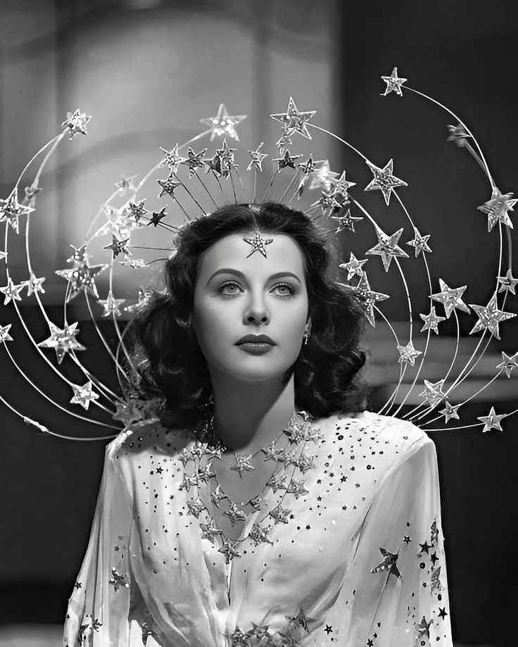 Uma foto em preto e branco da atriz Hedy Lamarr. Seus cabelos cacheados estão soltos e batem na altura dos ombros. Ela tem uma estrela brilhante colada no meio de sua testa. As estrelas também se fazem presentes em sua roupa, colar e em volta de sua cabeça em uma espécie de arco que, provavelmente, está conectado na parte de trás da vestimenta.