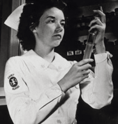 Foto em preto e branco da enfermeira Letitia Mumford. Letitia tem cabelos escuros em um comprimento médio, está usando seu uniforme de enfermeira e olha atentamente para a seringa que está segurando com ambas as mãos.