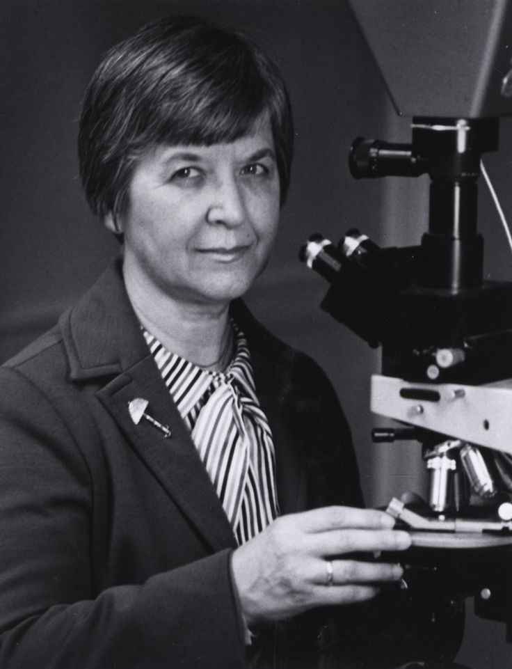 Foto em preto e branco da cientista Stephanie Kwolek. Stephanie possui cabelos curtos e está usando um sobretudo escuro sob uma blusa listrada. Uma de suas mãos está tocando levemente o que parece ser um equipamento de laboratório.