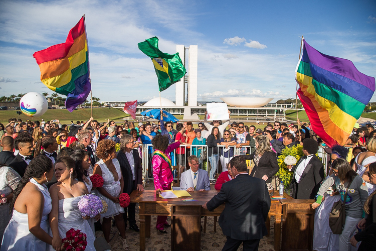 Fotografia de um casamento coletivo realizado numa área verde, diante do Congresso Nacional, em Brasília. No centro da imagem, há uma mesa de madeira com pessoas de terno sentadas, em frente a documentos que estão em cima da mesa. À frente da mesa, encontram-se vários casais com vestidos brancos e segurando buquês de flores estão reunidos para a cerimônia de casamento. À esquerda e à direita da imagem, uma multidão acompanha o evento e duas grandes bandeiras do arco-íris, ladeando essas pessoas, estão erguidas. Ao fundo, uma bandeira do Brasil também é levantada.