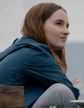 A imagem mostra uma jovem de perfil, sentada em um ambiente externo com clima nublado. Ela tem pele clara, cabelos lisos castanho-claros e está vestindo uma jaqueta azul escura e calça cinza.