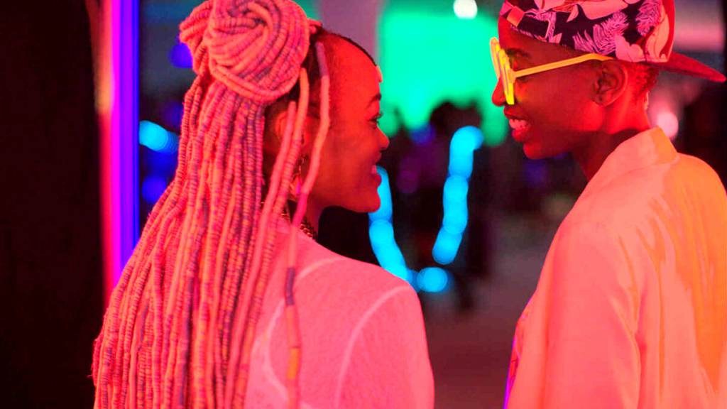 A imagem mostra duas mulheres negras se olhando com carinho em um ambiente iluminado por luzes neon coloridas. A pessoa à esquerda tem tranças longas e rosas, presas em parte por dois coques altos, e usa uma blusa branca translúcida. A pessoa à direita usa óculos amarelos fluorescentes, boné estampado virado para trás e uma camisa branca aberta, sorrindo para a outra. Ao fundo, luzes azuis e verdes.