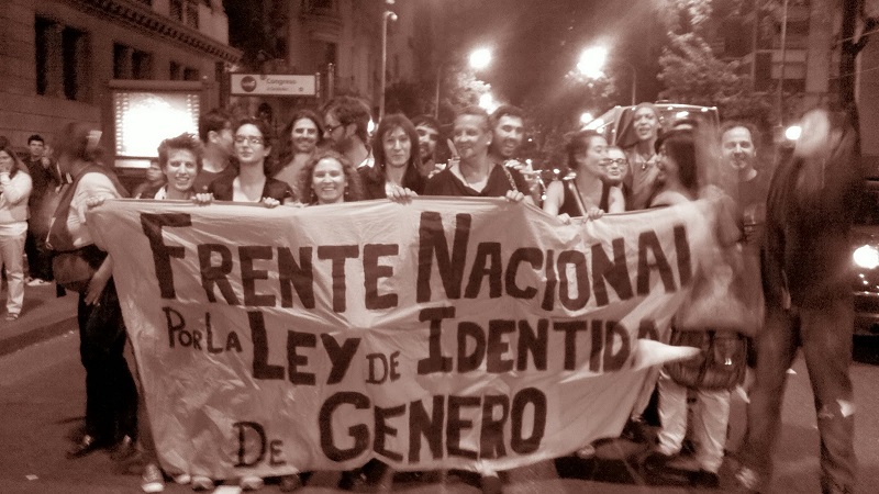 A imagem é composta por um grupo plural de manifestantes, em uma rua urbana iluminada por postes, no meio da noite. Em conjunto, ês manifestantes seguram uma grande faixa branca, com os dizeres "Frente Nacional Por La Ley de Identidad de Género", escrito à mão. Ês manifestantes estão sorrindo para a foto, demonstrando um momento de união e força coletiva. A foto é colorida em tons de sépia, trazendo uma sensação histórica para o visual.