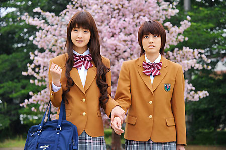 Aqui temos os dois protagonistas da série japonesa IS: Otoko demo onna demo nai sei, de mãos dadas e de costas para uma árvore de cerejeira. Do lado esquerdo está Miwako Aihara, sorridente, de cabelos castanhos longos e carregando uma bolsa azul escura em um dos braços. A direita temos Haru Hoshino, de cabelos também castanhos, porém curtos, apresentando uma expressão desconcertada ao encarar algo a sua frente. Ambos estão vestidos com o que aparenta ser seus uniformes escolares, consistentes em blazers marrons por cima de blusas se gola branca, gravatas vermelhas com listras brancas e as partes debaixo (possivelmente uma saia e uma calça) xadrez em tons de azul.