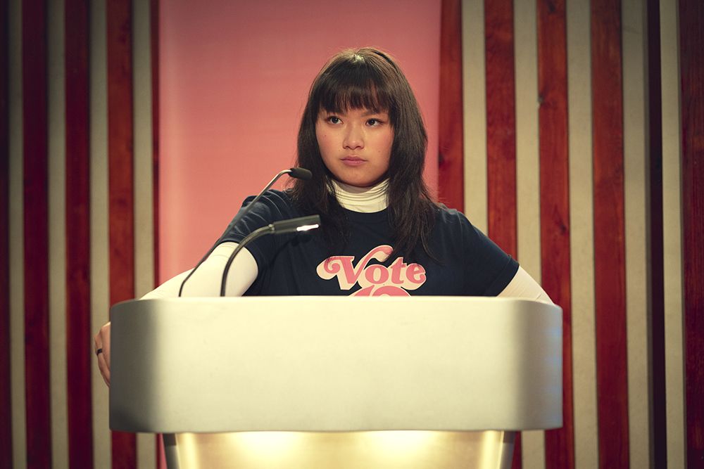 Na imagem, há uma jovem de expressão séria em pé atrás de um púlpito. Ela tem cabelo liso e franja, e veste uma camiseta azul-marinho com a palavra 'Vote' em rosa sobre uma blusa de manga longa branca. À frente dela, há dois microfones. O fundo é composto por painéis verticais de madeira em tons claros e escuros, com uma faixa rosa central.