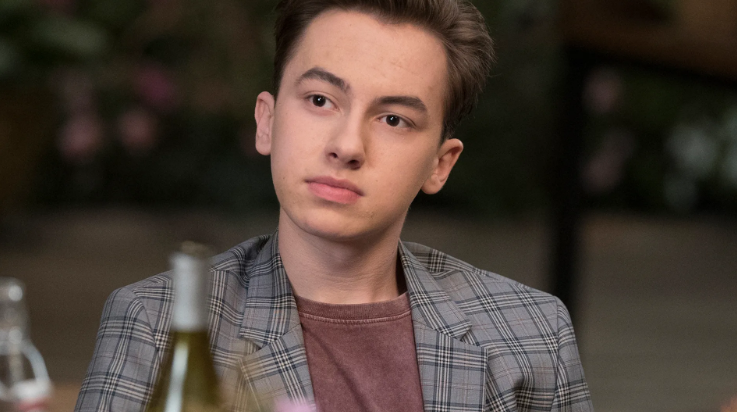 A imagem mostra um adolescente de pele branca com cabelo castanho escuro penteado para o lado. Ele veste uma camiseta marrom por baixo de um blazer xadrez em tons de cinza e está sentado, olhando levemente para o lado com uma expressão neutra ou pensativa.