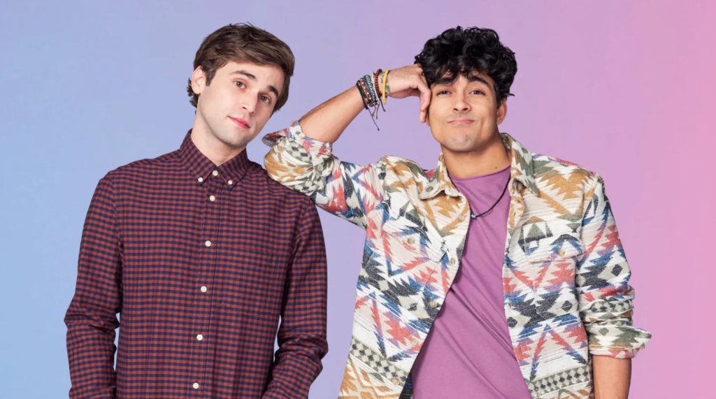 A imagem mostra dois jovens em pé, lado a lado, posando para a câmera contra um fundo em degradê azul e rosa. À esquerda, está um rapaz de pele branca, cabelo castanho curto e liso, usando uma camisa de botões xadrez em tons de vermelho e azul escuro. Ele tem uma expressão suave e um leve sorriso. À direita, está um rapaz de pele marrom, cabelo preto e encaracolado. Ele veste uma camiseta roxa e uma jaqueta estampada com padrões geométricos coloridos. Seu braço direito está levantado, com o cotovelo apoiado no ombro do outro rapaz, em um gesto descontraído. Ele usa várias pulseiras e sorri de leve.