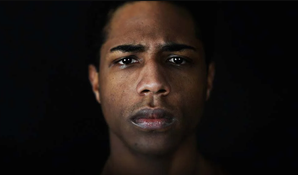 A imagem mostra um jovem negro de cabelos curtos pretos e um sinal no queixo, com o cenho franzido e uma expressão séria. Atrás dele está um fundo preto.