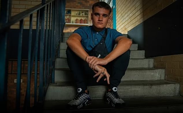 Na imagem, há um jovem sentado em uma escada de concreto. Ele tem cabelo curto, expressão séria e está vestindo uma camisa polo azul, calça escura e tênis pretos com branco. Usa uma pochete preta atravessada no peito e exibe um anel dourado em um dos dedos. O ambiente tem paredes de tijolos claros e um corrimão azul.