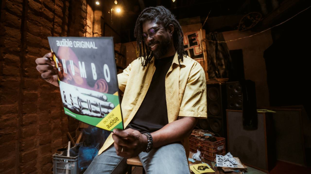Foto de Rafael Porcidônio, criador do podcast Chumbo & Soul, segurando o pôster do projeto com um sorriso no rosto. Ele é um homem negro retinto, com óculos e tranças na altura do ombro.O pôster é formado por duas ilustrações: em cima, de um homem negro, com black power e rosto comprimido. Embaixo, a de uma mão branca esticada, dando a sensação de que o homem está sendo calado por ela. O texto, em letras brancas, diz: audible original, CHUMBO & SOUL.Rafael está sentado em um banquinho em um pequeno quarto com caixas, materiais de som e discos.