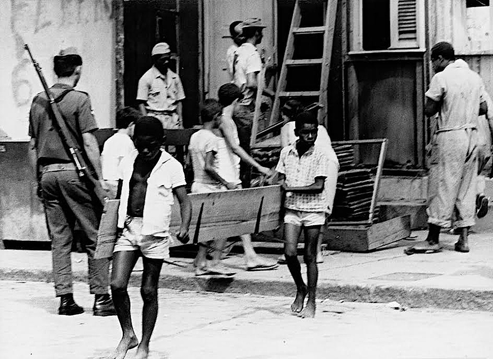 Duas crianças negras carregam uma tábua para fora de casa, enquanto outras observam o lugar de longe e policiais observam o lugar. A foto, em preto e branco, foi tirada durante a remoção de favelas no Rio de Janeiro.