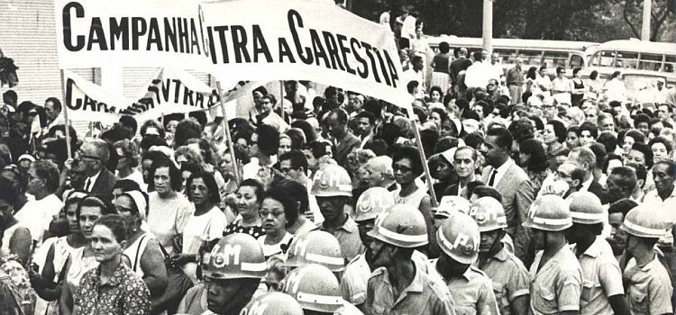 Imagem de um protesto. Homens e mulheres civis estão no centro, marchando e carregando cartazes (em um deles, é possível ler “Campanha Contra a Carestia”), enquanto são rodeados por militares.