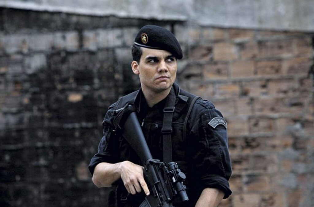 Na imagem, há um homem vestindo uniforme policial preto e boina preta com emblema, segurando um fuzil com as duas mãos. Ele tem expressão séria e olhar voltado para o lado, aparentando estar em alerta. No uniforme, há um patch no braço. Ao fundo, um muro de tijolos e cimento em ambiente urbano.