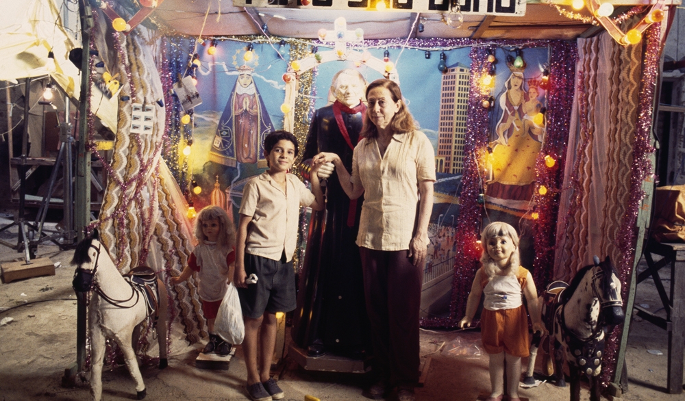 A imagem é uma cena colorida mostrando uma mulher idosa e um menino de mãos dadas, posando diante de um altar decorado com luzes coloridas, fitas e imagens religiosas ao fundo. Entre eles, há uma estátua vestindo batina preta com faixa vermelha. Ao lado, dois bonecos de tamanho quase real, loiros e com roupas coloridas, estão próximos a cavalos de brinquedo. O cenário remete a uma feira de rua popular.