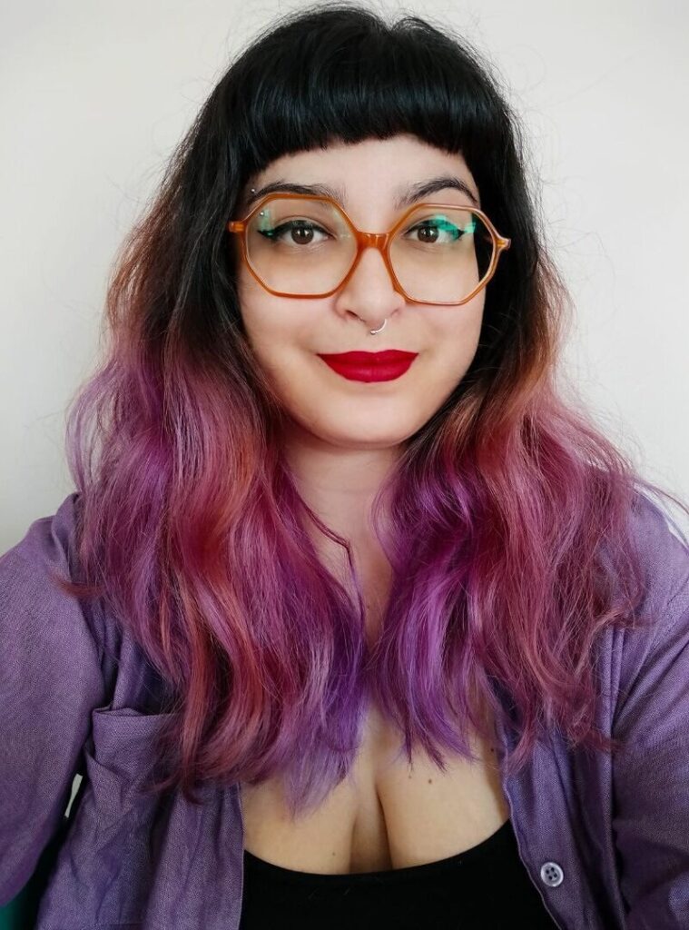 Mulher de pele branca, olhos castanhos e cabelos compridos nas cores castanho, roxo e rosa, com um pequeno sorriso de boca fechada. Ela usa uma blusa preta com uma camisa roxa por cima,  óculos arredondados de cor laranja e armação hexagonal, piercing no septo, batom vermelho e delineador preto.