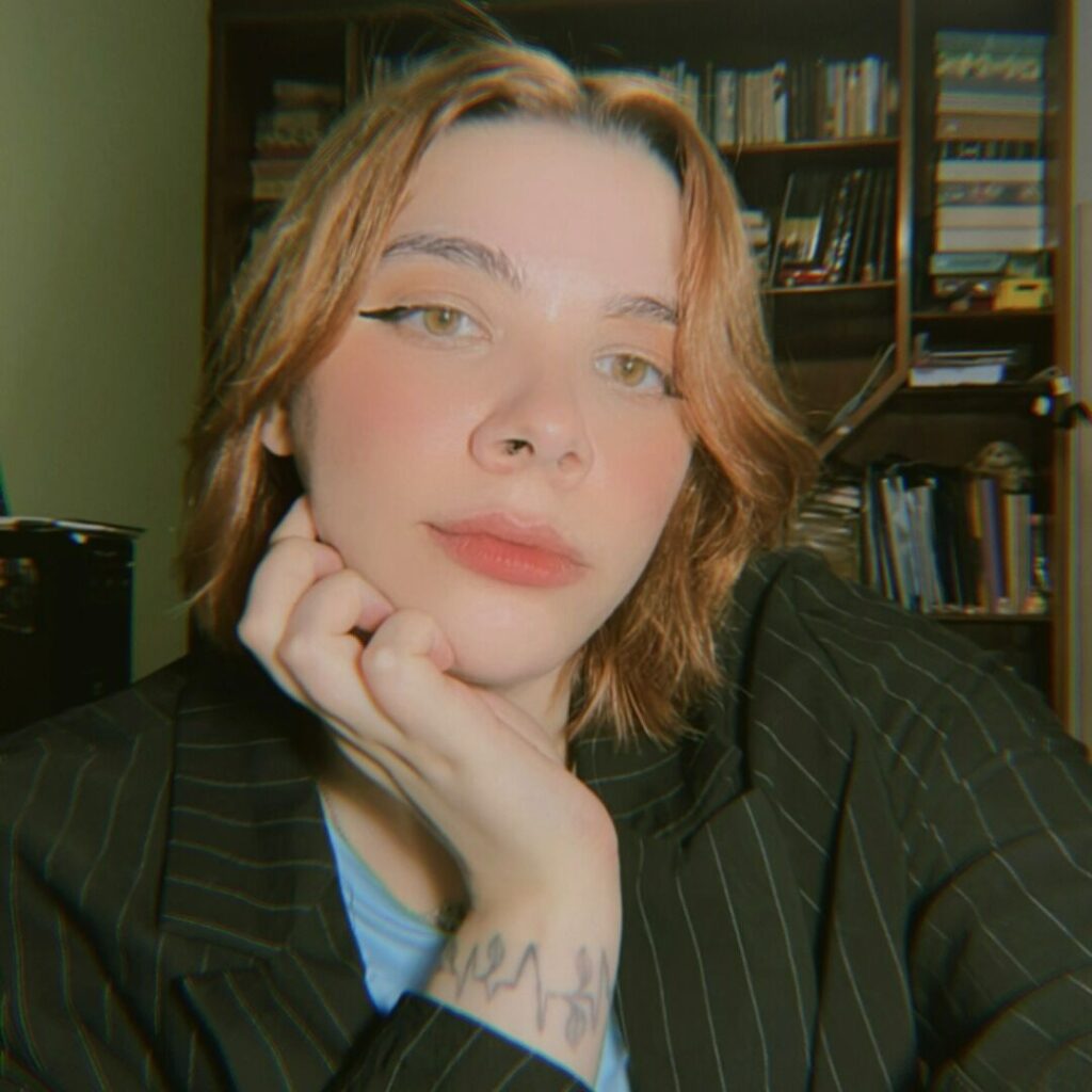 Pessoa de pele branca e cabelos curtos e lisos de tom castanho-alaranjado. Ela está com a mão apoiada no queixo e tem uma tatuagem à mostra no pulso. Usa um blazer risca de giz e uma blusa azul-clara por baixo.