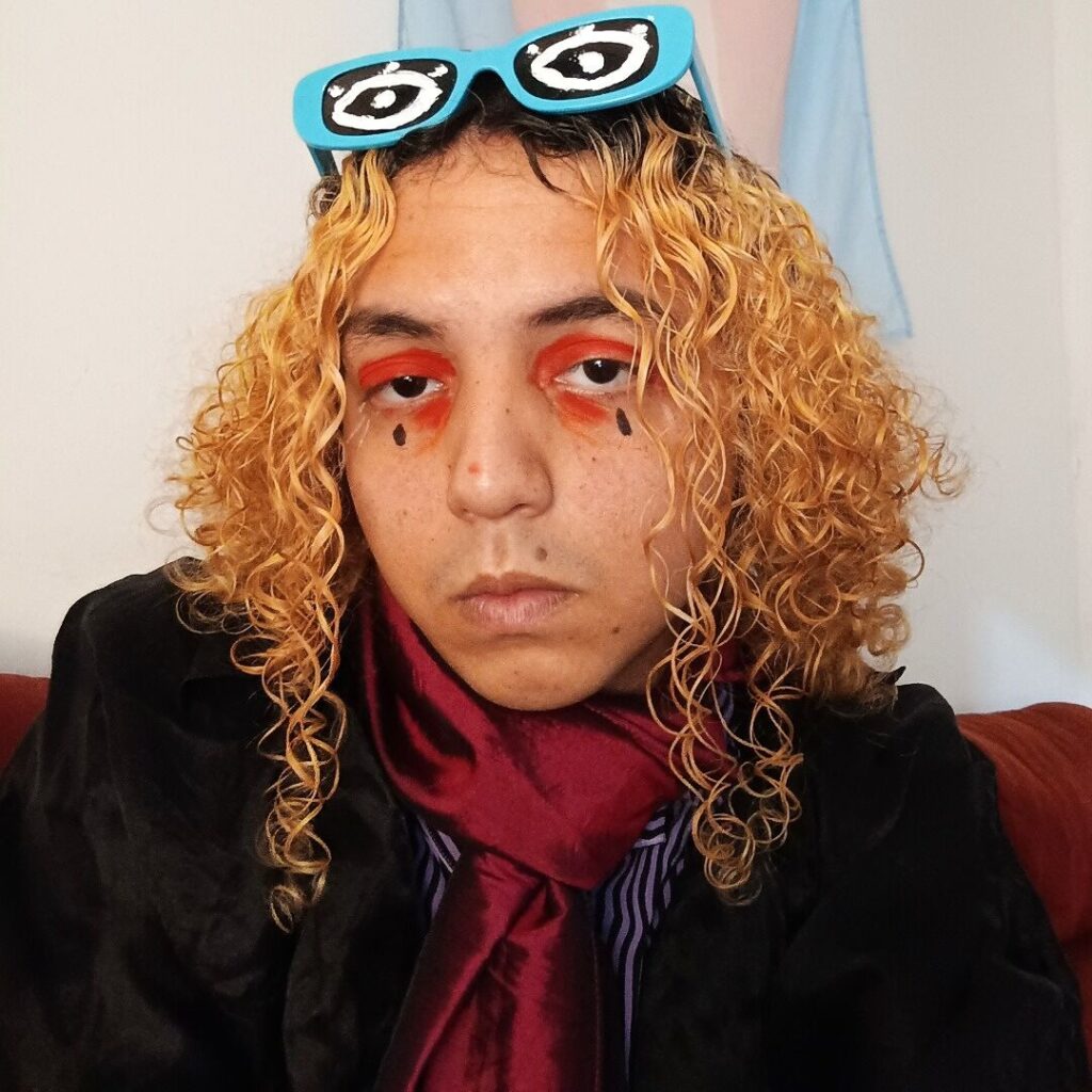 Pessoa de pele branca e cabelos ruivos curtos e cacheados, com sombra vermelho-alaranjada nos olhos e uma expressão séria no rosto, No topo da cabeça, usa óculos azuis-claros com olhos brancos pintados nas lentes. Usa um blazer preto, uma camisa listrada por baixo e uma echarpe vermelha de seda.