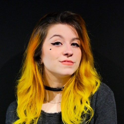 Mulher de pele branca, olhos azuis e cabelos compridos amarelos exibindo um pequeno sorriso de boca fechada. Ela usa delineador preto, gargantilha preta, camisa cinza e uma pequena estrela preta na bochecha. O fundo da foto é todo preto.